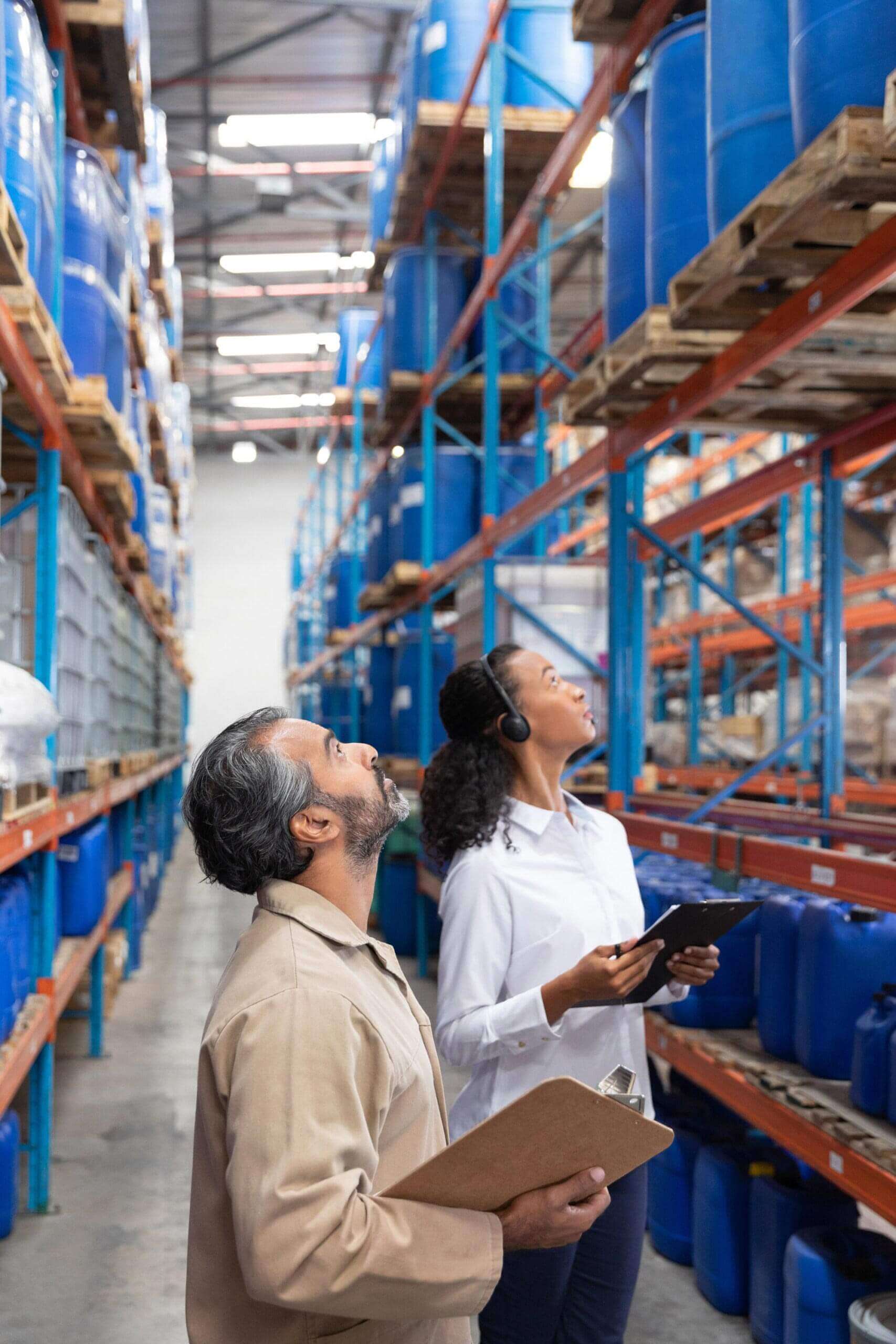 Large warehouse with shelves stacked with blue barrels; man and woman looking up holding a tablet and clipboard